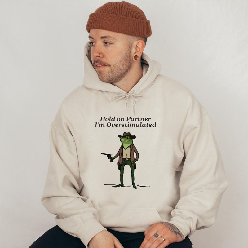 Young man wearing a brown beanie and a sand-colored hoodie featuring a graphic of a green frog dressed as a cowboy holding a revolver with text reading Hold on Partner I'm Overstimulated
