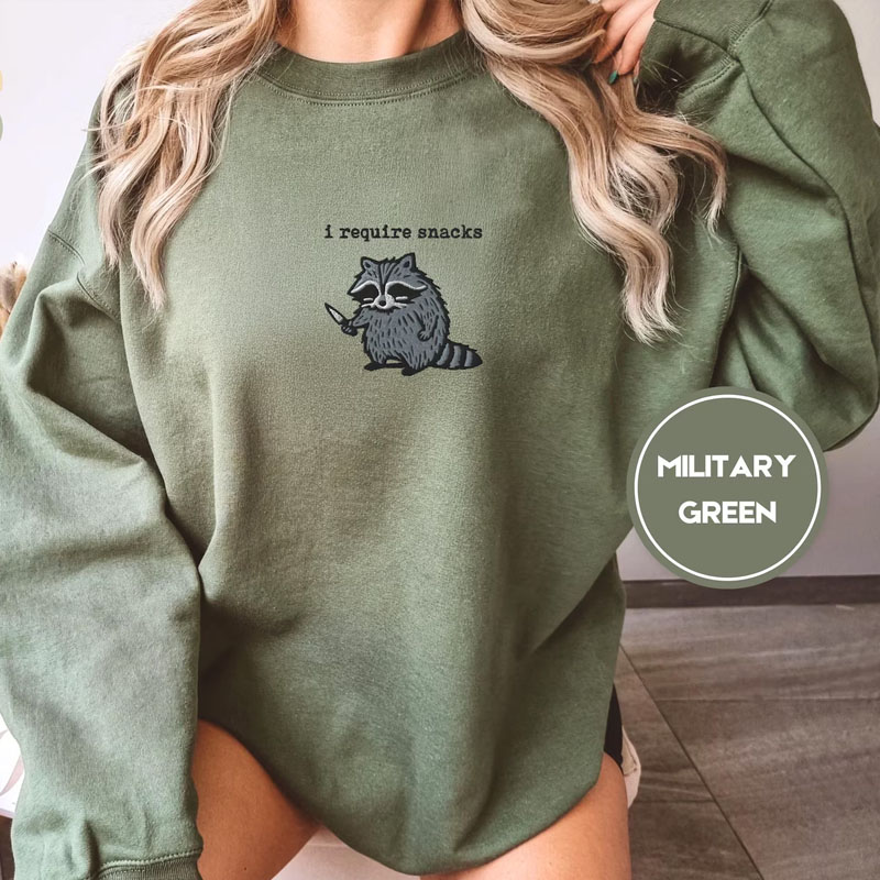 Woman wearing an oversized military green Gildan 18000 sweatshirt with high-quality embroidery of a feral raccoon holding a knife and text reading i require snacks