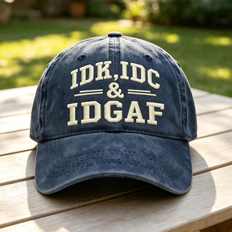 Vintage navy blue washed cotton baseball cap embroidered with beige text reading IDK IDC AND IDGAF resting on a wooden picnic table outdoors